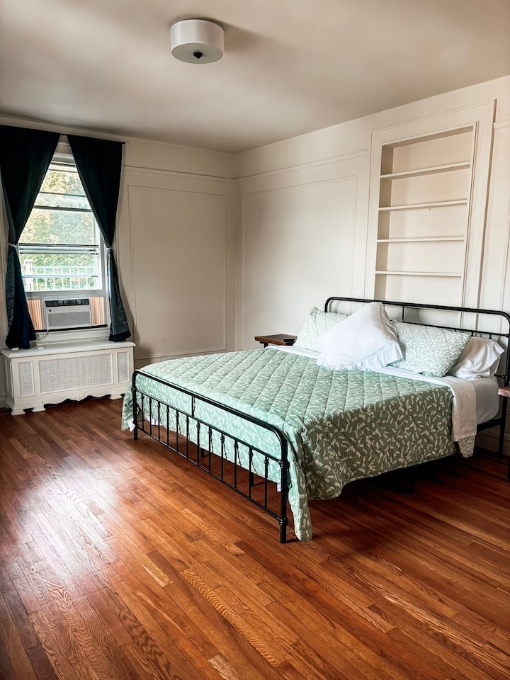 A serene master bedroom featuring a minimalist iron bed frame with sage green quilted bedding. The room is painted in soft off-white with traditional picture frame molding on the walls, creating a timeless editorial feel under bright, natural Saratoga Springs sunlight.