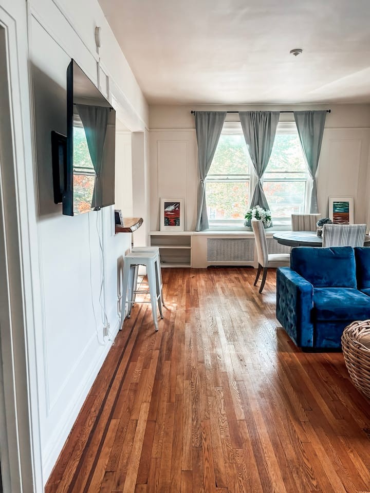 A spacious and elegant living room in a boutique Saratoga Springs apartment featuring wide-plank wood flooring, large windows with soft gray drapes, and high-end modern furniture in rich navy and warm sand tones. The lighting is soft and natural, emphasizing the minimalist and sophisticated architectural details.